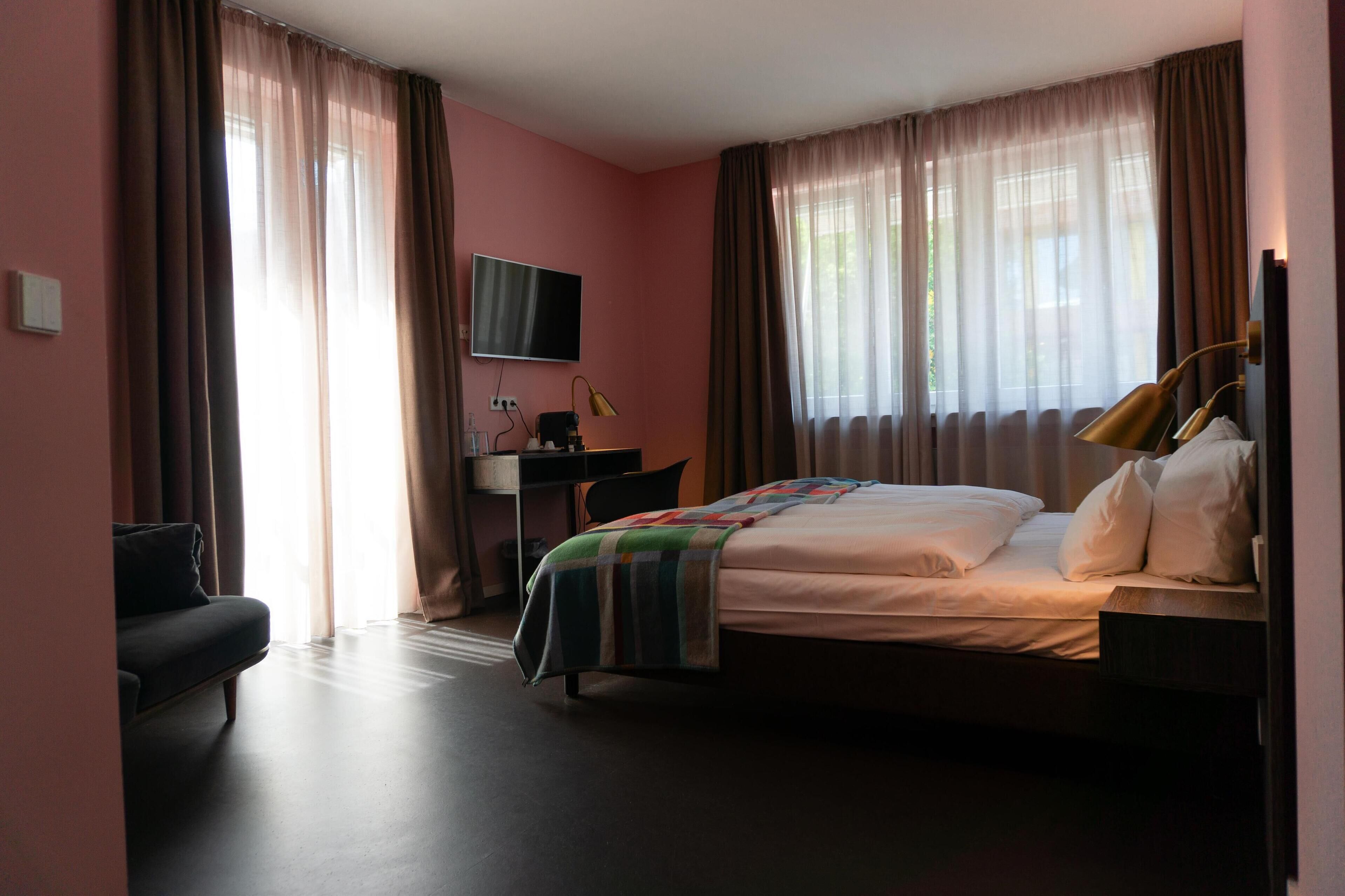 A hotel room with a bed, a desk, a chair, a TV, and large windows with curtains.