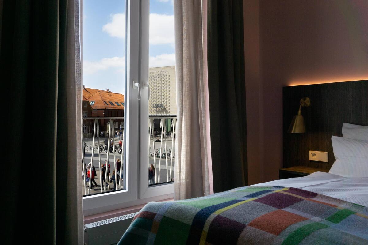 A hotel room with a large window, a bed with a colorful blanket, a nightstand with a lamp, and a view of a street with people and bicycles.