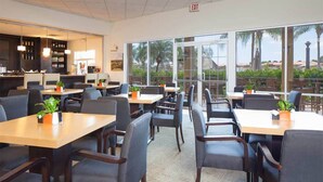 A restaurant with tables and chairs, a bar area, and large windows offering a view of palm trees and buildings. - Encantada Resort (Kissimmee)