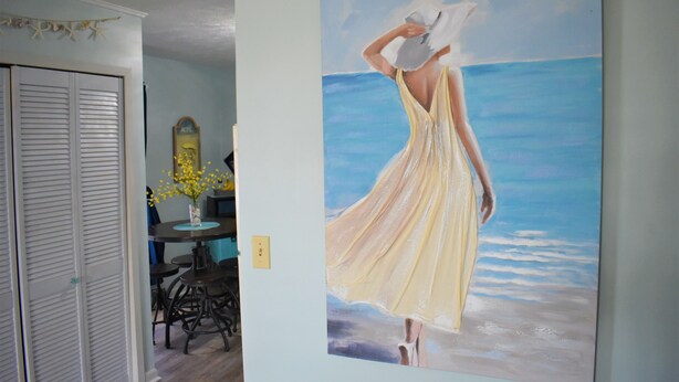 Una habitación con un cuadro de una mujer con vestido amarillo frente a una ventana con vista al océano.
