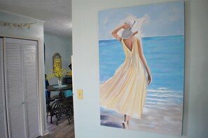 A room with a painting of a woman in a yellow dress by a window overlooking the ocean.