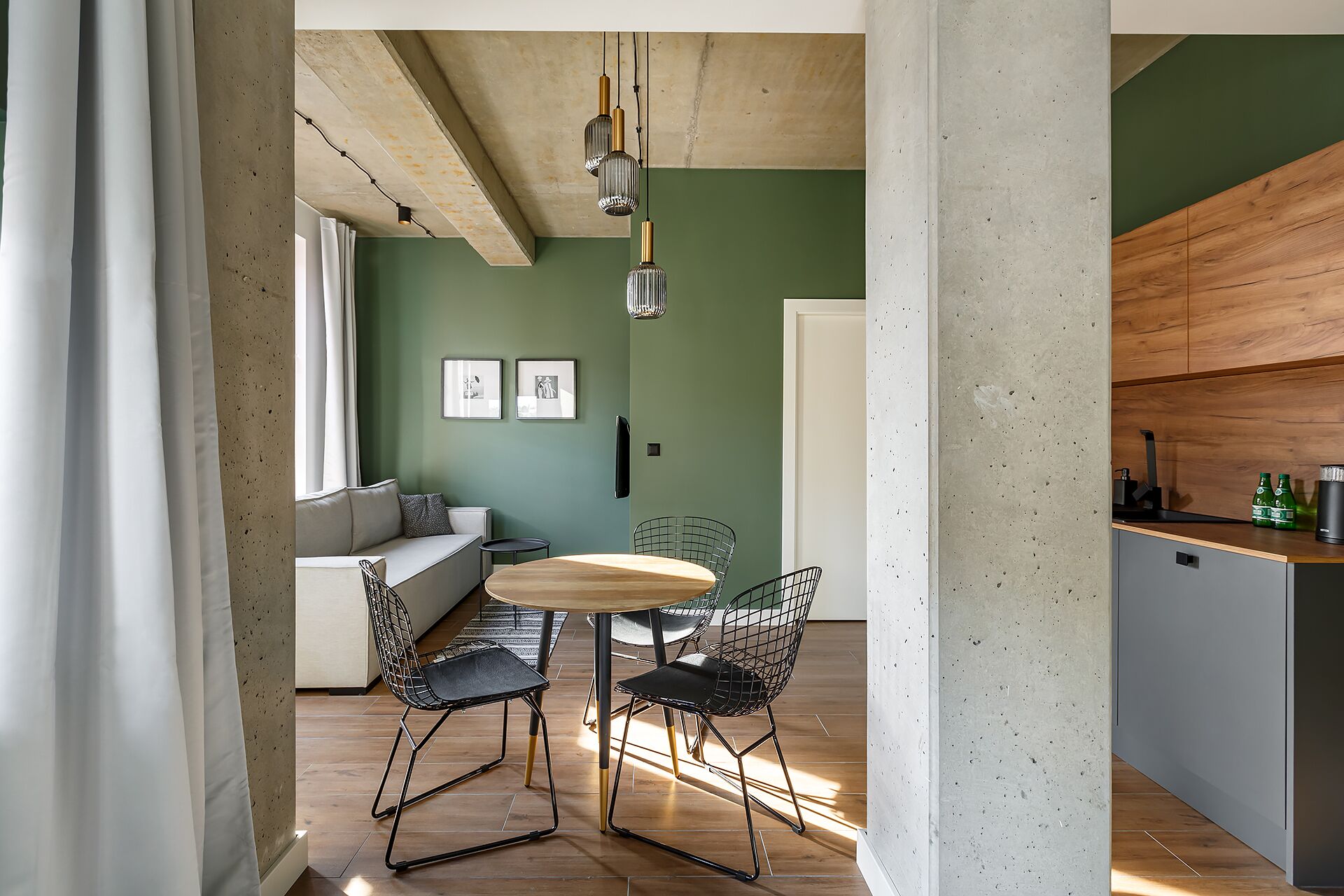 A modern living space with a dining area featuring a round wooden table and wire mesh chairs, a sofa, and a green accent wall.