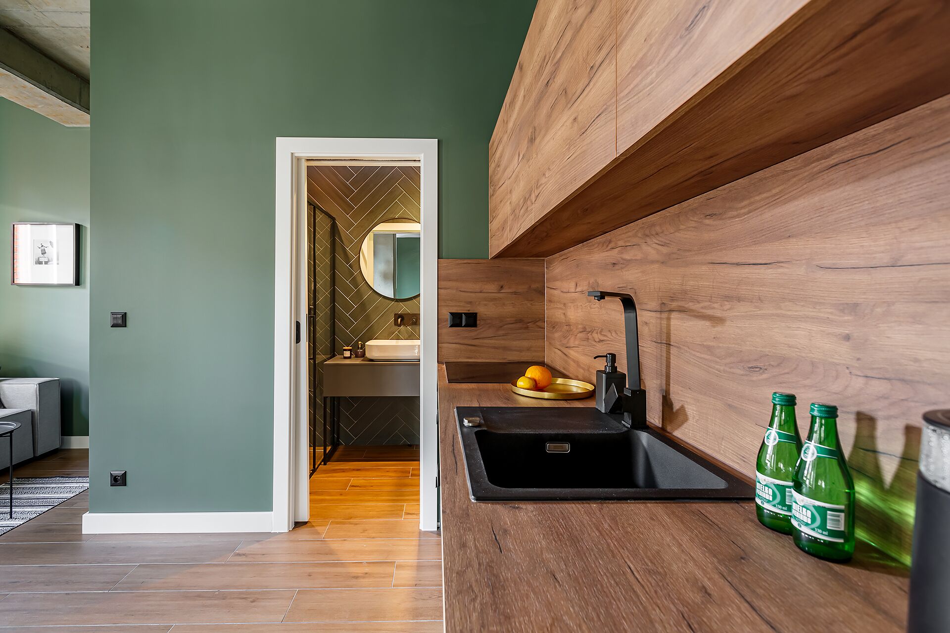 A modern bathroom with a wooden vanity, a sink, and a mirror. There are two green bottles on the counter.