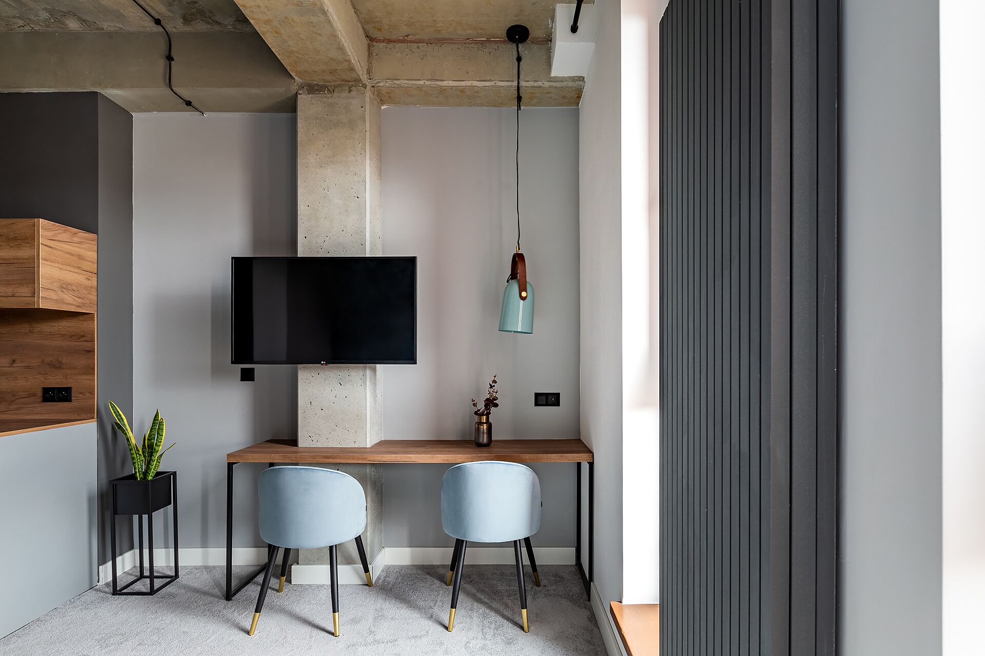 Une pièce moderne avec un bureau en bois, deux chaises, un téléviseur fixé sur un pilier en béton et un luminaire suspendu.