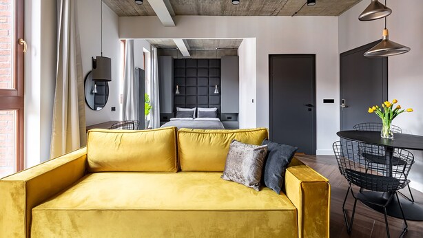 A modern living space with a yellow sofa, a dining table with chairs, and a kitchen area in the background.