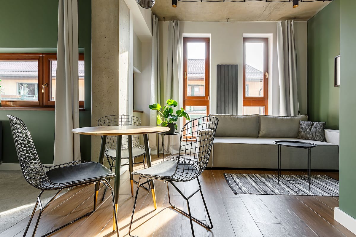 Una sala de estar moderna con piso de madera, una mesa redonda, dos sillas de estructura metálica, un sofá y una mesita auxiliar con una planta.