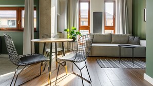 A modern living room with a wooden floor, a round table, two wireframe chairs, a sofa, and a small side table with a plant.
