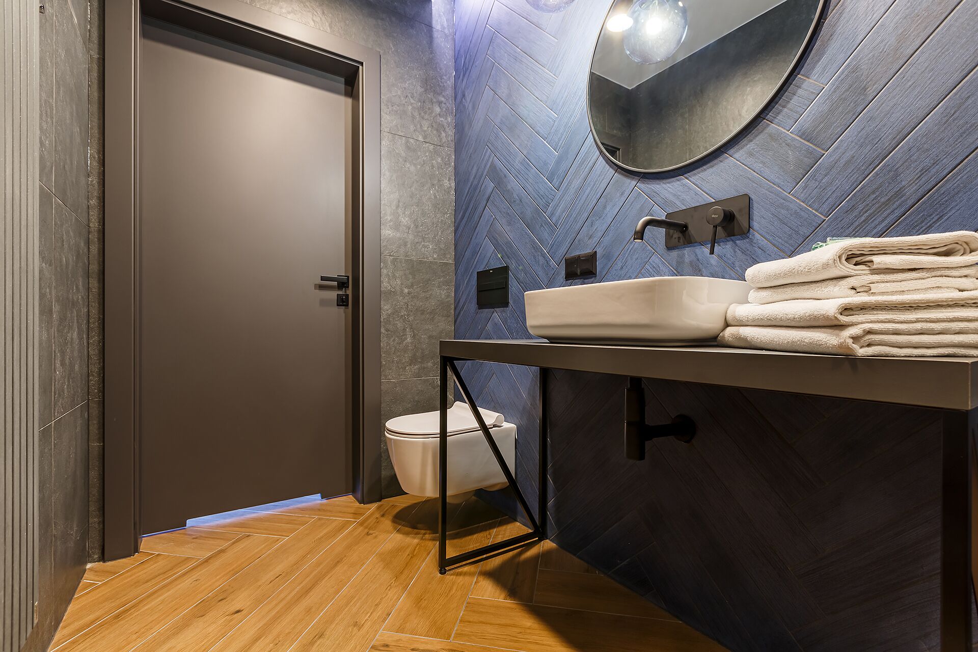 Une salle de bain moderne avec un plancher en bois, une porte sombre, un lavabo avec un miroir et des toilettes.
