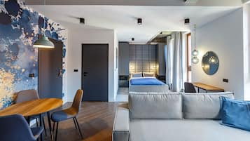 A modern hotel room with a sofa, a dining area with a table and chairs, a bed with a headboard, and a wall with a unique blue and white design.