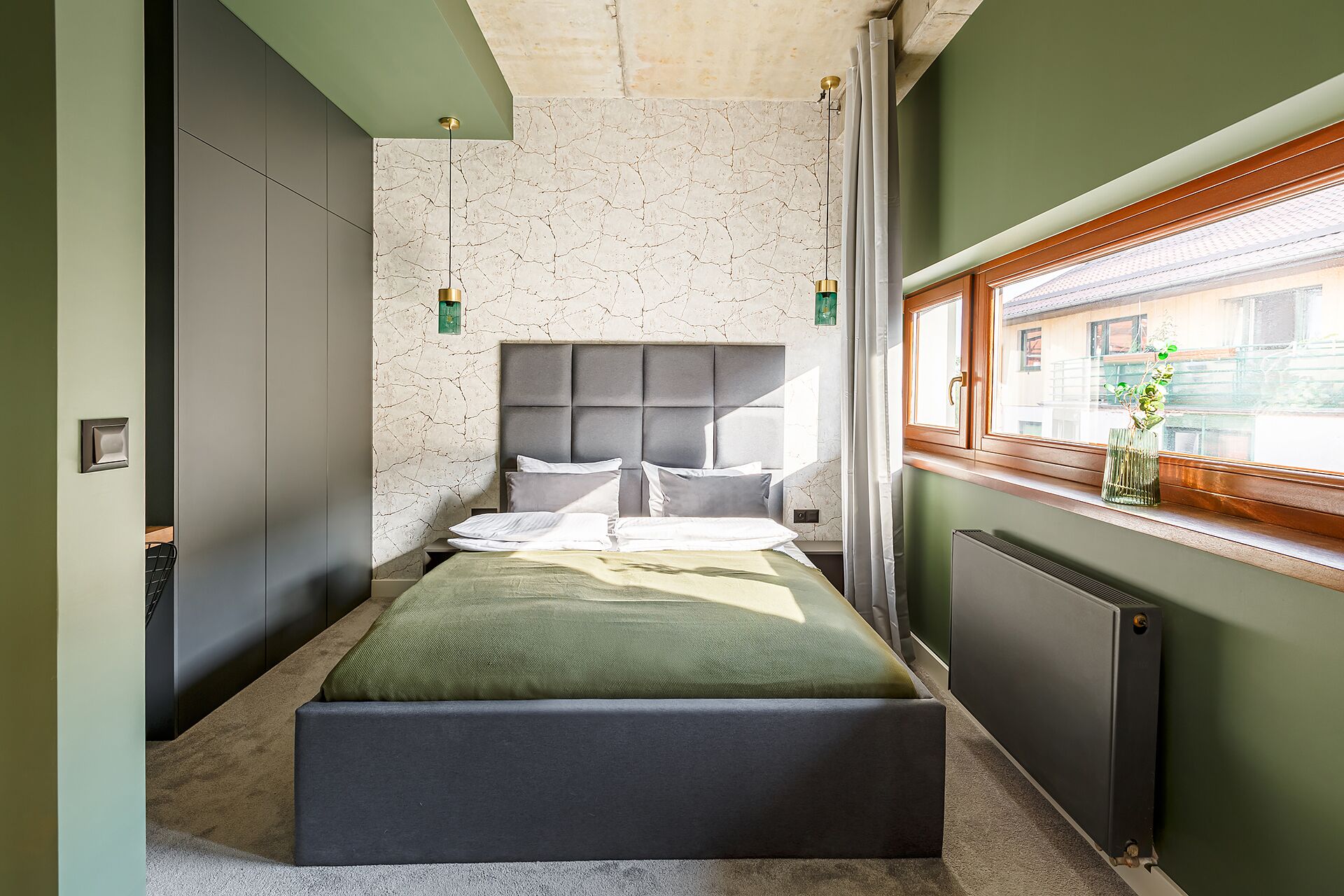 A modern bedroom with a large bed, a headboard, a radiator, and a window with a view of buildings.