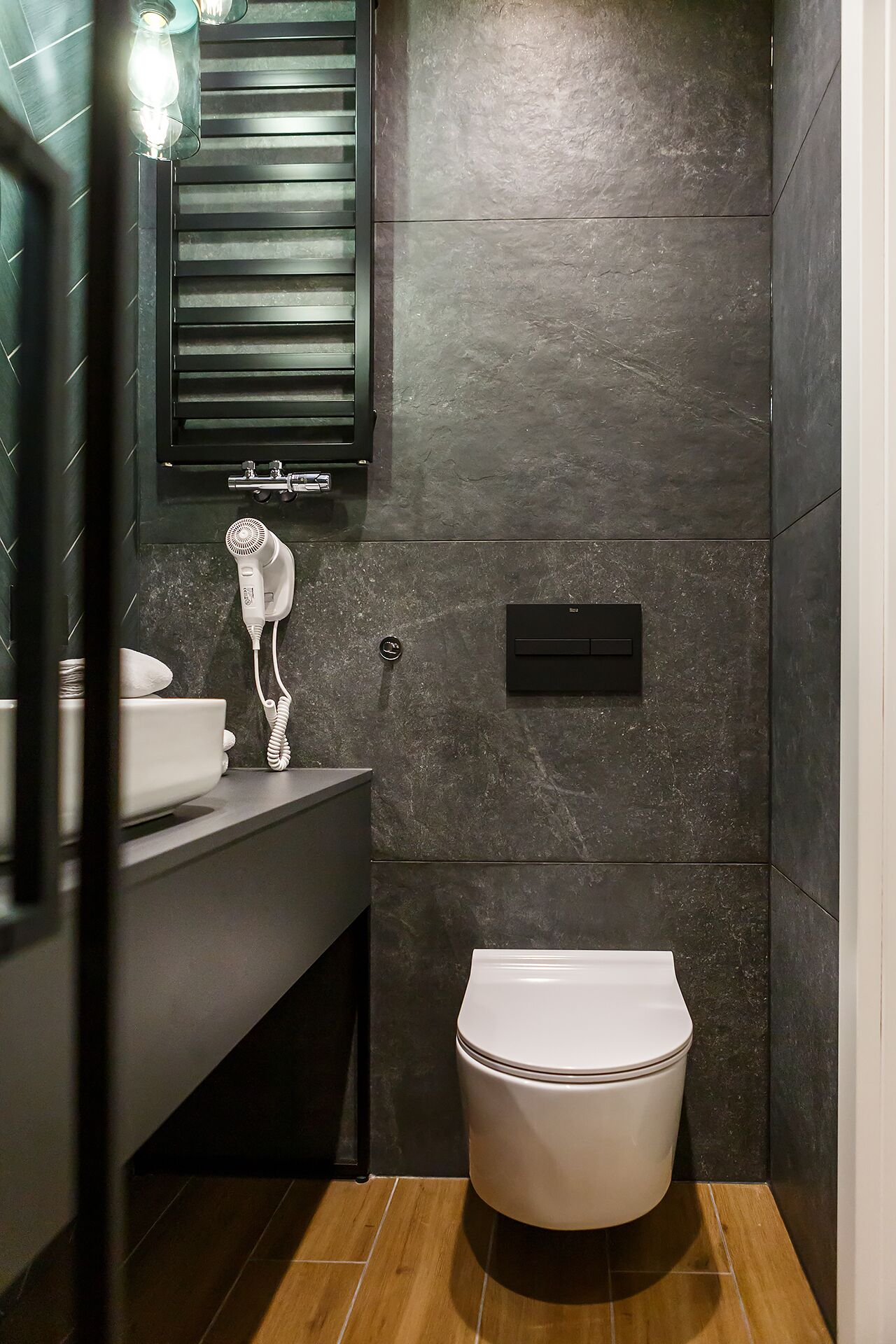 A modern bathroom with a dark tiled wall, a white toilet, a sink with a towel, and a wall-mounted heater.