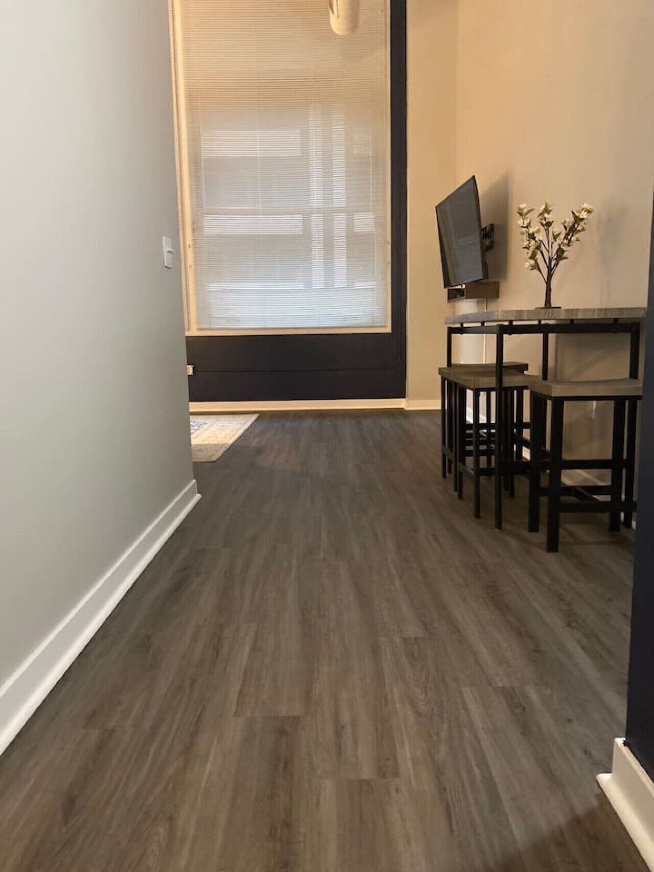 A room with a dark wood floor, a TV on a stand, a small table with chairs, and a window with blinds.