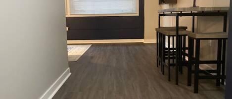 A room with a dark wood floor, a TV on a stand, a small table with chairs, and a window with blinds.