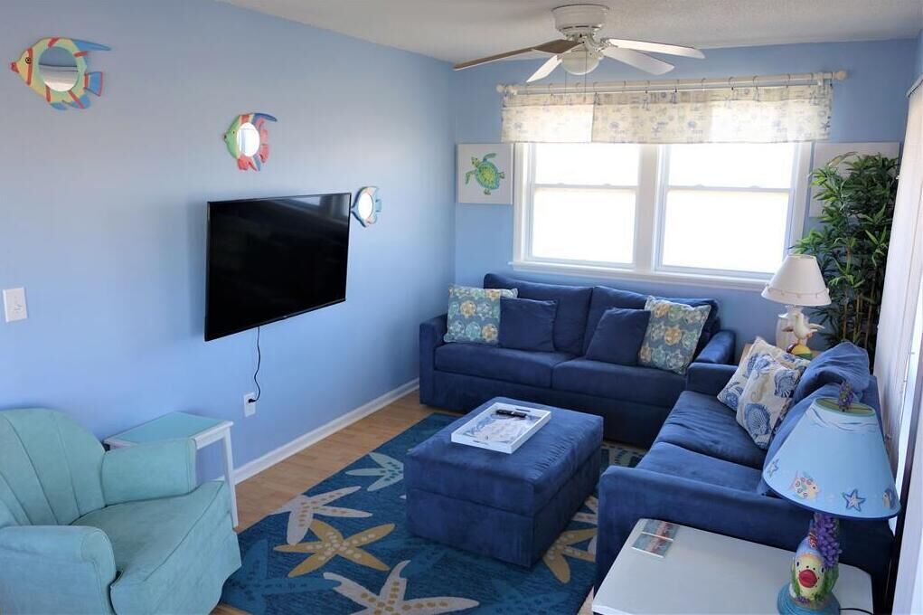 Una sala de estar con un sofá azul, un televisor, un ventilador de techo y una ventana con cortina.