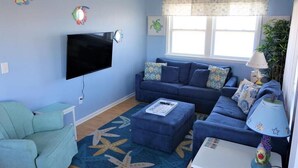 Una sala de estar con un sofá azul, un televisor, un ventilador de techo y una ventana con cortina.