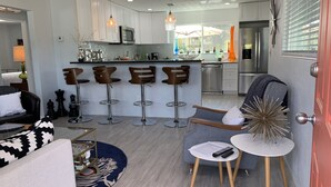 A modern kitchen with white cabinets, a central island, and bar stools. There is a dining area with a gray sofa and a small round table. - Mid Century home close to beaches and downtown Dunedin. 1 block from the Trail. (Clearwater)
