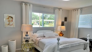 A bedroom with a bed, two bedside lamps, a window with curtains, a wall-mounted picture, and a small table with a lamp. - Mid Century home close to beaches and downtown Dunedin. 1 block from the Trail. (Clearwater)