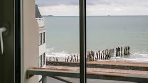 Une vue depuis une fenêtre donnant sur une zone côtière, avec une digue en bois et un bâtiment doté d’un balcon.