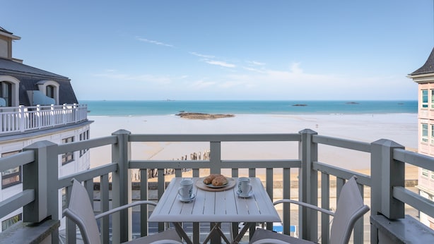 Un balcon avec une table dressée pour deux personnes, donnant sur une plage sous un ciel dégagé.
