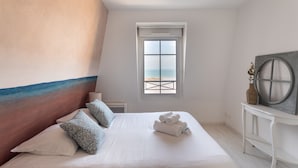 Une chambre avec un grand lit, une vue sur la plage par la fenêtre et une commode blanche surmontée d’un vase.