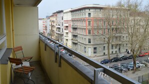 A balcony with two wooden chairs, a yellow wall, and a view of multi-story buildings with parked cars and a tree. - Vacation apartment in the heart of Berlin ( City West ) (Berlin)