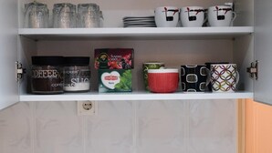 En køkkenhylde med forskellige ting, herunder kaffedåser, en kasse Suma kaffe og dekorative krus.