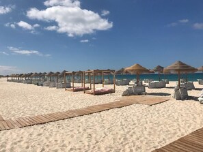 Een strand met rijen ligbedden en rieten parasols, houten looppaden en een helder uitzicht op de oceaan.