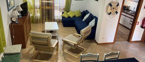 A living room with a sofa, chairs, a small table, and a TV. There is a kitchen area visible through an open door.