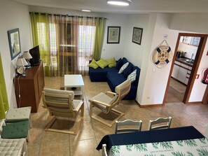 Una sala de estar con un sofá, sillas, una mesita y un televisor. Se ve un área de cocina a través de una puerta abierta.
