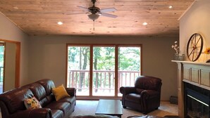 A living room with a ceiling fan, a fireplace, and a large window overlooking a deck. - Amazing Experiences Await Your Group - Family or Friends (Bella Vista)