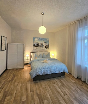 A bedroom with a bed, bedside lamps, a ceiling light, a painting, and a wardrobe.