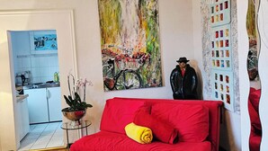 Un salón con un sofá rojo, un cuadro de una bicicleta y una cocina visible al fondo.
