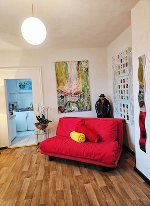Ein Wohnzimmer mit einem roten Sofa, einem Gemälde eines Fahrrads und einer im Hintergrund sichtbaren Küche.