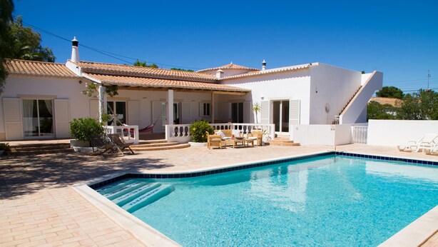 A swimming pool with a tiled deck, a covered patio area with outdoor furniture, and a white building with a tiled roof.