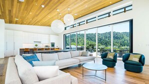 Amplia sala de estar con un sofá grande, dos sillones y una mesa de centro. Se ve una cocina al fondo, y hay ventanas amplias que ofrecen vistas a un área boscosa.