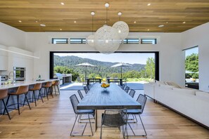A modern kitchen with a long dining table, wooden flooring, and a view of the mountains.