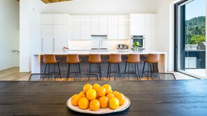 Una cocina moderna con armarios blancos, una isla central con taburetes de bar y una mesa de comedor de madera con un plato de naranjas.
