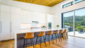 Una cocina moderna con una isla grande, taburetes de bar y vistas a una ladera boscosa.
