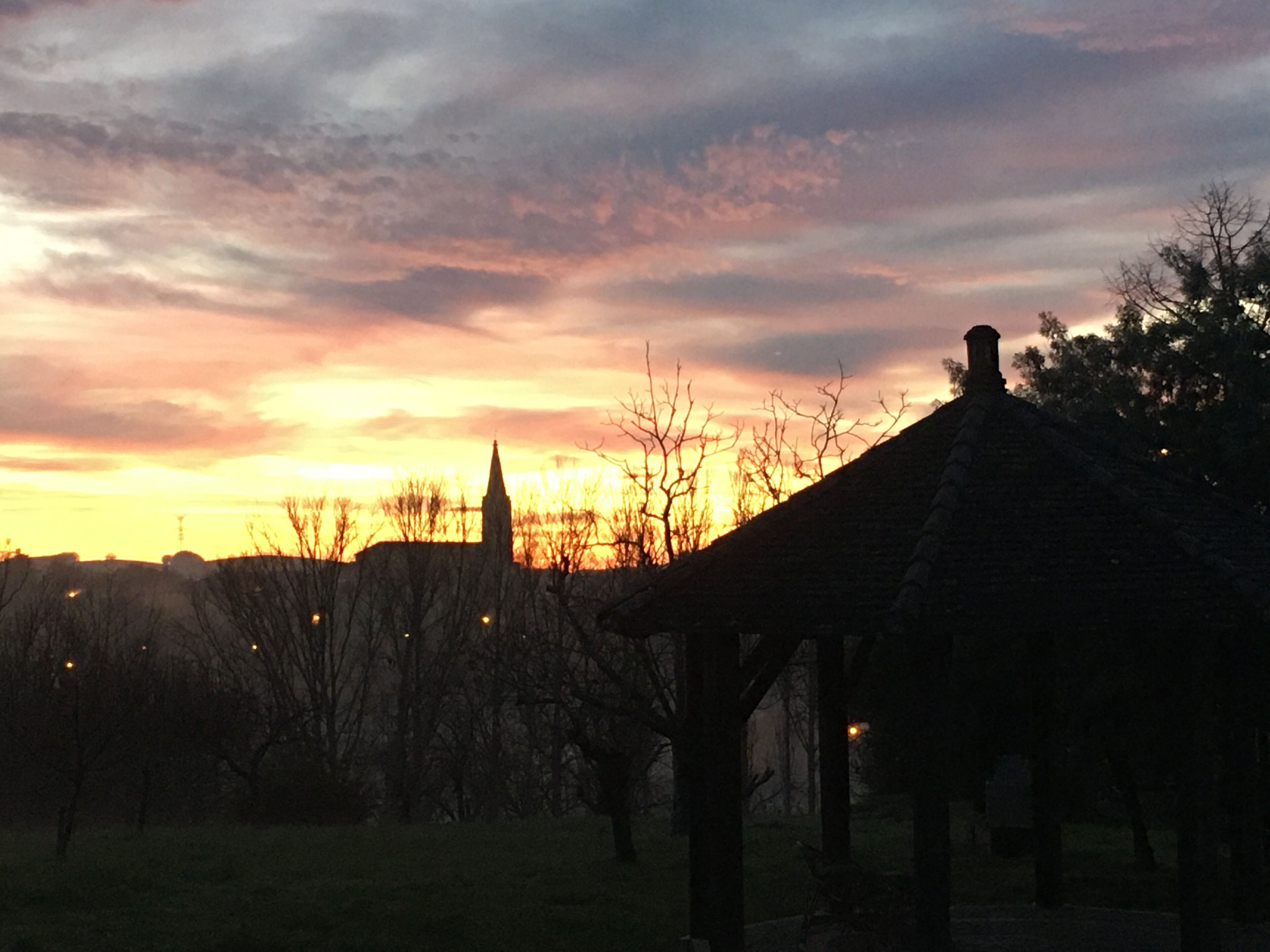 Un coucher de soleil sur un paysage rural, avec le clocher d’une église se détachant sur le ciel, entouré d’arbres et d’un champ d’herbe.