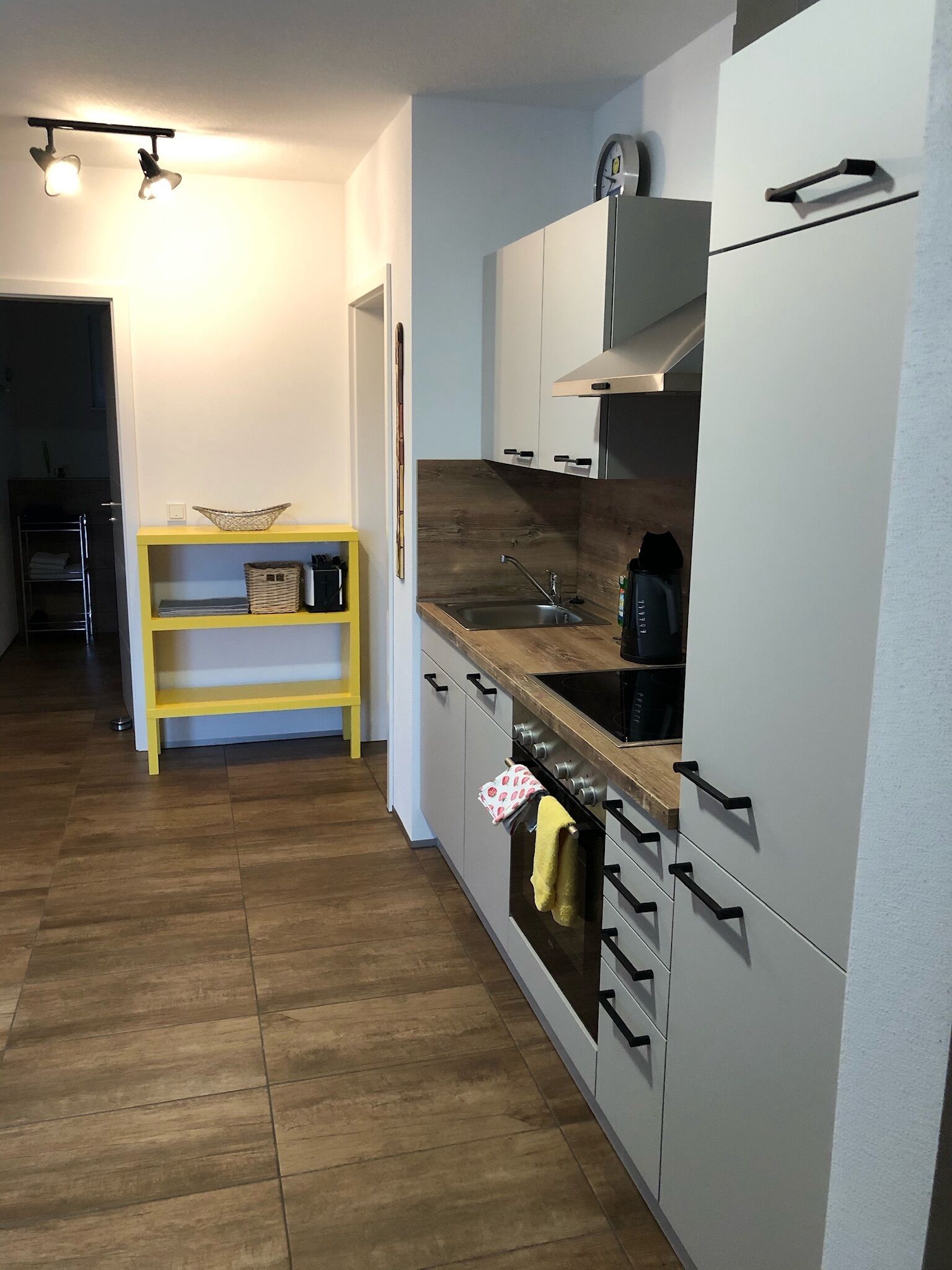 Una cocina moderna con encimera de madera, pileta de acero inoxidable y muebles de cocina blancos. Se observa una estantería amarilla con cestas y un reloj en la pared.