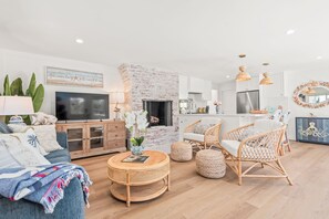 Un salon avec un téléviseur, une cheminée, des meubles en osier et une cuisine à l’arrière-plan.