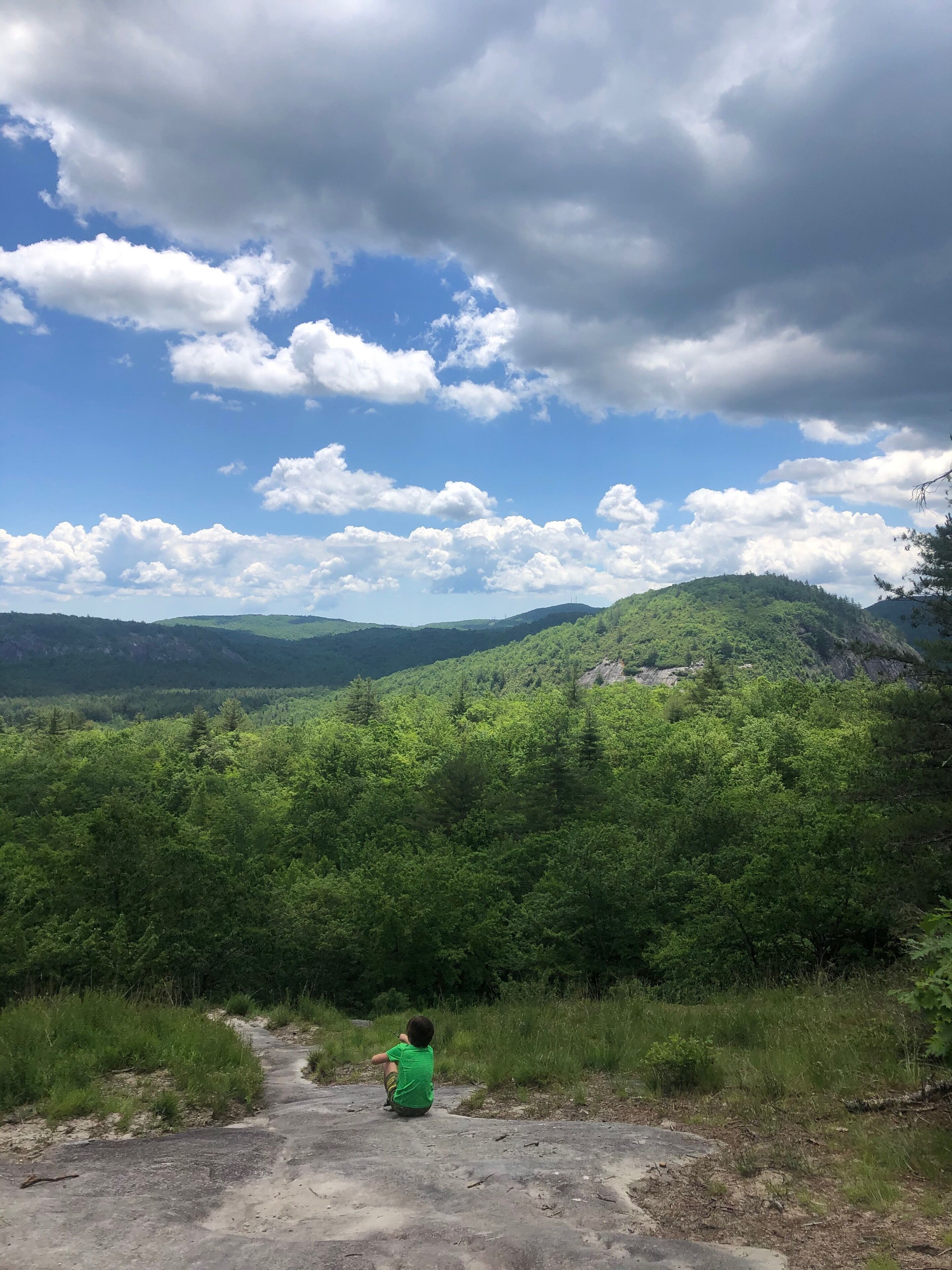 A person sitting on a concrete path surrounded by lush green hills and a partly cloudy sky.