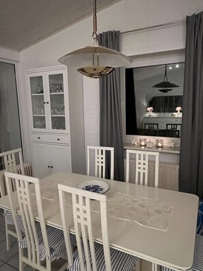 Un'area pranzo con tavolo e sedie bianche, un mobile contenitore con vetri, un lampadario e una finestra con tende.
