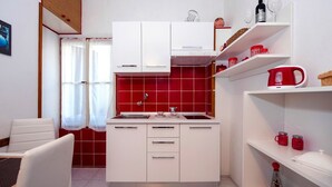 A compact kitchen with white cabinets, a red tiled backsplash, and a sink. There are shelves with red mugs and a kettle. - STELA - One Bedroom Apartment (Rovinj)