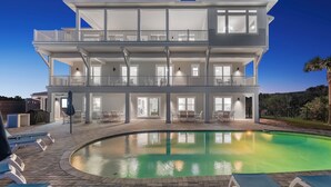 Una casa frente a la playa de dos pisos con piscina, mobiliario exterior y un patio cubierto.