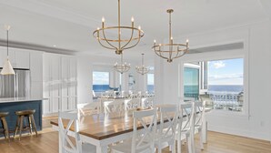 Een ruime eetruimte met een houten tafel en witte stoelen, grote ramen met uitzicht op de oceaan, en elegante kroonluchters.