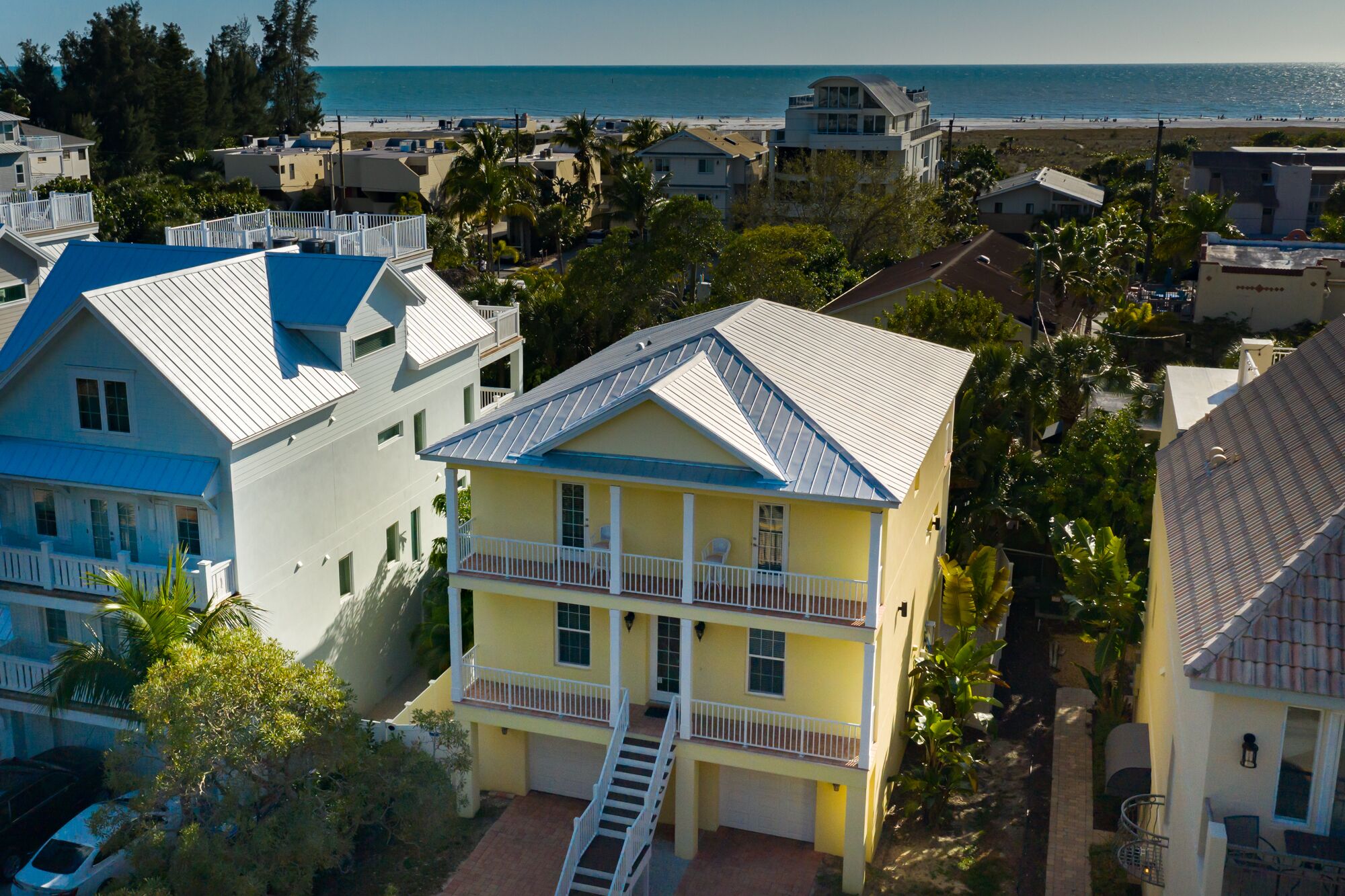 A multi-story yellow building with a balcony, surrounded by other residential buildings and a beach in the background.