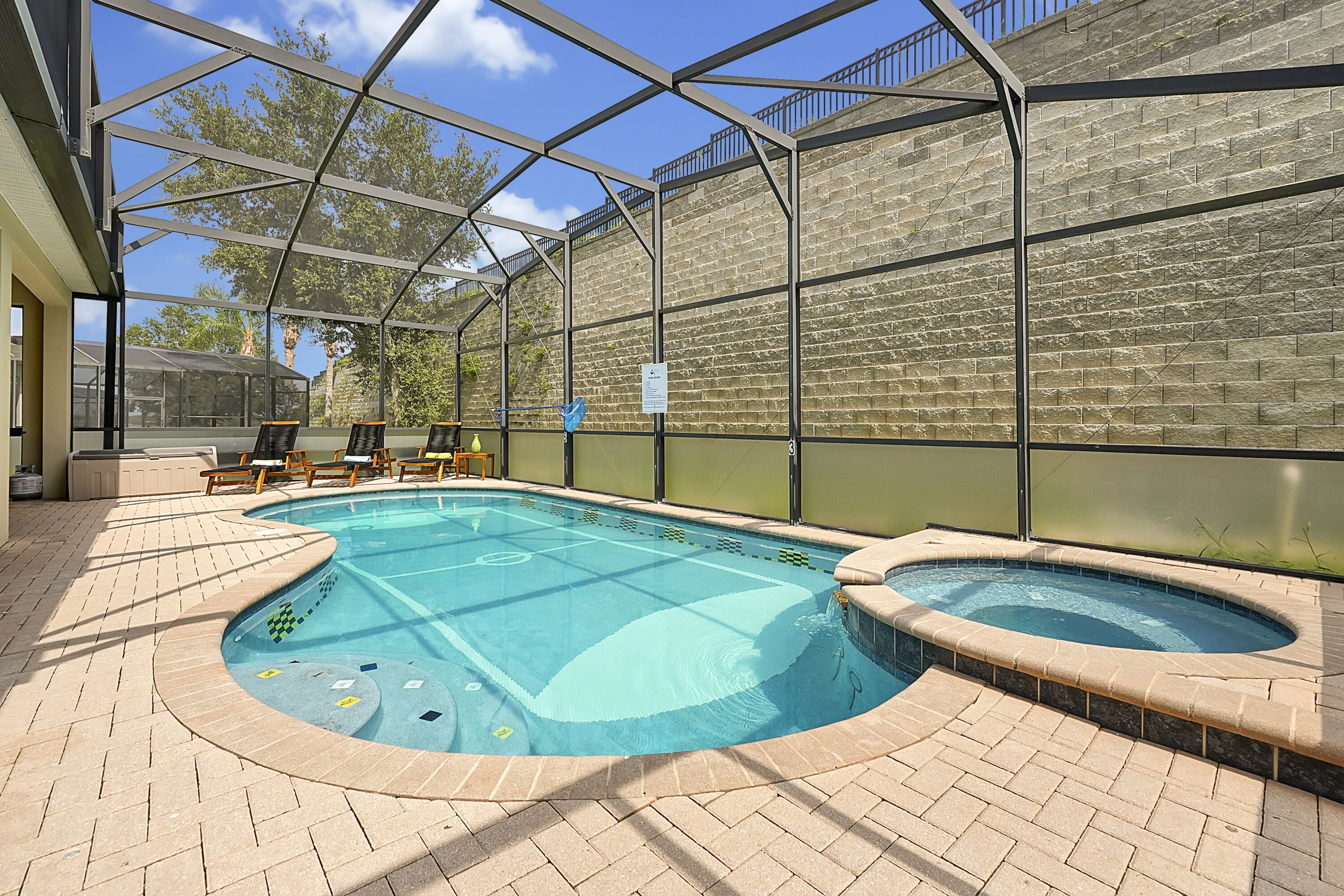 A covered swimming pool with a hot tub, lounge chairs, and a brick patio.