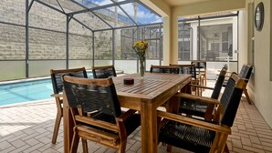 A covered patio with a pool, wooden table and chairs, and a vase of yellow flowers.