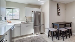 A modern kitchen with white cabinets, a stainless steel refrigerator, a dining table with four chairs, and a gray rug on the floor.